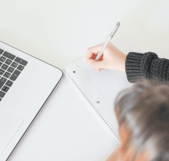 Woman writing notes next to laptop in asychronous webinar. Credit J Kelly